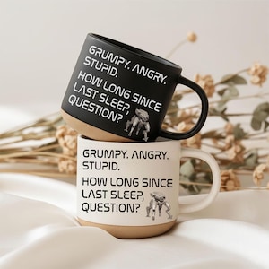 Könnte beinhalten: Zwei Keramikbecher, einer schwarz und einer cremefarben, übereinander gestapelt. Beide Becher tragen den Text: "GRUMPY. ANGRY. STUPID. HOW LONG SINCE LAST SLEEP, QUESTION?" Jeder Becher hat eine kleine Roboter-Grafik.