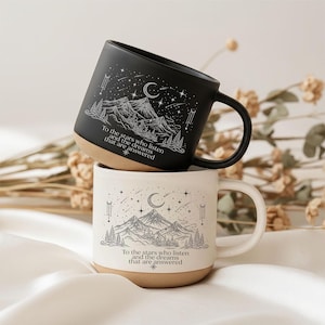 Könnte beinhalten: Ein schwarzer Keramikbecher und ein weißer Keramikbecher, beide mit einer Illustration von Berglandschaft, Mond und Sternen sowie dem Text: To the stars who listen and the dreams that are answered. Beide Becher haben einen naturfarbenen Boden.