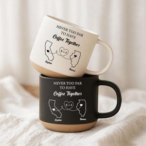 Può includere: Due tazze in ceramica impilate verticalmente. La tazza superiore è color crema con il testo nero NEVER TOO FAR TO HAVE Coffee Together, che mostra i contorni della California e dell'Illinois collegati da una linea tratteggiata a un cuore.