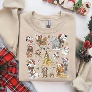 Könnte beinhalten: Beiges Sweatshirt mit festlichem Design verschiedener Hunderassen, die Weihnachtsmützen und Schals tragen. Das Design umfasst dekorative Elemente wie Sterne und Zweige, die ein weihnachtliches Thema schaffen.