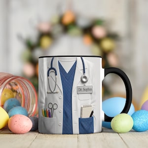 Könnte beinhalten: Weiße Keramiktasse mit einem Design eines Arztkittels, einem blauen Oberteil, einem Stethoskop und dem Namen "Dr. Sophia". Die Tasse hat einen schwarzen Henkel und Rand. Bunte Ostereier sind im Hintergrund.