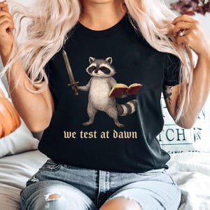 Pode incluir: Camiseta preta com uma ilustração de um guaxinim segurando uma espada e um livro. O guaxinim está em pé, com uma espada na mão direita e um livro na esquerda. O texto "we test at dawn" está impresso abaixo.