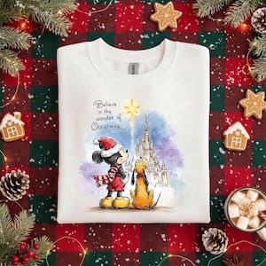 Könnte beinhalten: Weißes Sweatshirt mit Rundhalsausschnitt und Weihnachtsgrafik. Das Design zeigt Micky Maus und Pluto vor einem Schloss mit dem Text "Believe in the wonder of Christmas". Das Sweatshirt ist mit Lebkuchen und Tannenzapfen verziert.
