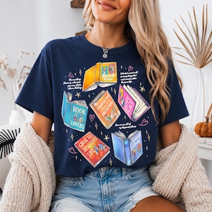 Könnte beinhalten: Ein dunkelblaues Kurzarm-T-Shirt mit einem Grafikdruck von mehreren aufgeschlagenen Büchern mit bunten Einbänden und Titeln wie Beach Read, People We Meet On Vacation, Happy Place, Book Lovers, Great Big Beautiful Life und Funny Story.