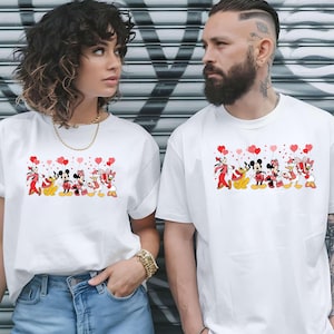 Pu&ograve; includere: T-shirt bianche con una grafica colorata di personaggi Disney che tengono in mano palloncini a forma di cuore. I personaggi includono Topolino, Minnie, Paperino e Paperina. Le magliette sono indossate da un uomo e una donna.
