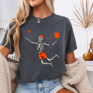 Könnte beinhalten: Ein anthrazitfarbenes T-Shirt mit einer Grafik eines Skeletts, das eine orangefarbene Baseballkappe und orangefarbene Shorts trägt. Das Skelett springt, um einen orangefarbenen Pickleball mit einem Schläger zu schlagen, mit dem Text well, shit...
