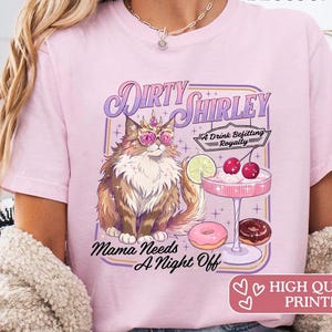 Könnte beinhalten: Ein hellrosa T-Shirt mit einer Grafik einer flauschigen Katze, die eine Krone und eine rosa Sonnenbrille trägt. Der Text lautet Dirty Shirley, A Drink Befitting Royalty und Mama Needs A Night Off. Ein Cocktailglas mit Kirschen, einer Limettenscheibe und zwei Donuts sind abgebildet.