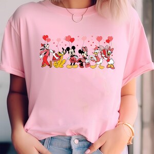 Pu&ograve; includere: T-shirt rosa chiaro con una grafica dei personaggi Disney, tra cui Pippo, Pluto, Topolino, Minnie, Paperino e Paperina, ognuno con palloncini a forma di cuore. I personaggi indossano i loro abiti classici. La maglietta &egrave; di colore rosa unito.