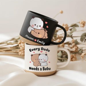 May include: A black ceramic mug with a white and brown bear illustration and the text Mason Emily, stacked on a speckled white ceramic mug featuring a brown and white bear illustration with the text Every Dudu Needs a Bubu in black font.