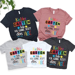 Könnte beinhalten: Fünf T-Shirts in verschiedenen Farben, darunter Marineblau, Rosa, Grau, Weiß und Anthrazit. Jedes Shirt zeigt den Text "Kindergarten Ade Ich Lerne Jetzt das ABC" in einem farbenfrohen Blockbuchstaben-Design mit Sternakzent.