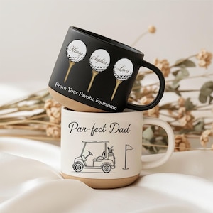 Könnte beinhalten: Zwei Keramikbecher, einer schwarz und einer weiß, übereinander gestapelt. Der schwarze Becher hat drei Golfball-Designs mit Namen und dem Text "From Your Favorite Foursome". Der weiße Becher zeigt einen Golfwagen und den Text "Par-fect Dad."