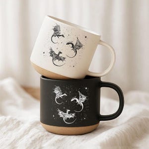 Könnte beinhalten: Ein Stapel aus zwei Keramikbechern mit Henkeln. Der obere Becher ist cremeweiß mit schwarzen Drachenillustrationen und Sternen. Der untere Becher ist mattschwarz mit weißen Drachenillustrationen und Sternen. Beide haben einen naturfarbenen Boden.