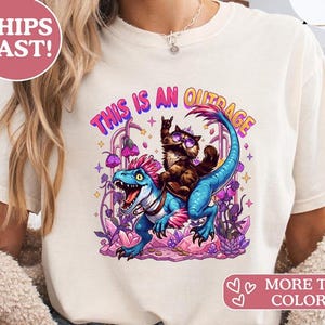 Könnte beinhalten: Cremefarbenes T-Shirt mit einer Cartoon-Illustration. Eine braune Katze mit Sonnenbrille reitet auf einem blauen Dinosaurier, darüber steht der Text "THIS IS AN OUTRAGE". Das Design enthält lila und rosa Akzente.