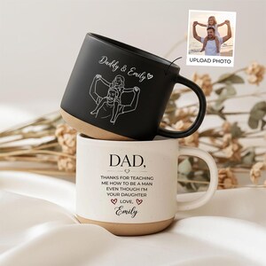 Könnte beinhalten: Ein mattschwarzer Becher mit einer weißen Strichzeichnung eines Elternteils, das ein Kind auf den Schultern trägt, und dem Text Daddy & Emily, gestapelt auf einem cremefarbenen Becher mit dem Text DAD, THANKS FOR TEACHING ME HOW TO BE A MAN EVEN THOUGH I'M YOUR DAUGHTER LOVE, Emily.