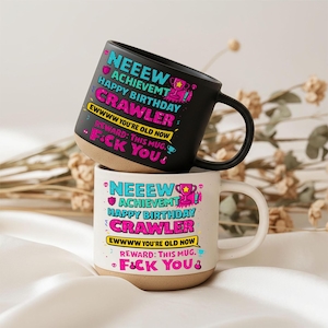 Könnte beinhalten: Zwei Keramikbecher, die vertikal gestapelt sind. Der obere Becher ist mattschwarz, der untere ist cremeweiß. Beide zeigen den identischen bunten Text: NEEEW ACHIEVEMT HAPPY BIRTHDAY CRAWLER EWWWW YOU'RE OLD NOW REWARD: THIS MUG. F*CK YOU mit Pokalsymbolen.