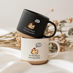 Könnte beinhalten: Ein mattschwarzer Keramikbecher gestapelt auf einem gesprenkelten weißen Keramikbecher. Beide Becher zeigen eine Pixel-Art-Grafik eines schlafenden Huhns mit einer Sprechblase mit ZZZ und dem Text I'm starting to feel exhausted.