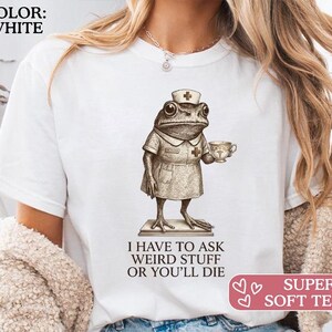 Könnte beinhalten: Weißes T-Shirt mit einer detaillierten Illustration einer Frosch-Krankenschwester, die eine Teetasse hält. Der Text "I HAVE TO ASK WEIRD STUFF OR YOU'LL DIE" steht darunter. Das Shirt hat auch den Text "SUPER SOFT TEES!"