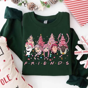 Könnte beinhalten: Dunkelgrünes Sweatshirt mit einer Grafik von Zeichentrickfiguren und Weihnachtsbäumen. Das Wort "FRIENDS" ist in Rosa gedruckt. Das Sweatshirt hat lange Ärmel mit dunkelgrünen Bündchen.