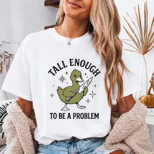 Könnte beinhalten: Ein weißes kurzärmeliges T-Shirt mit einer Grafik einer olivgrünen Ente, die ein großes Messer hält. Der Text über der Ente lautet TALL ENOUGH und der Text darunter TO BE A PROBLEM, ergänzt durch kleine schwarze Sternsymbole.