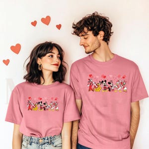 Pu&ograve; includere: Due persone indossano magliette rosa abbinate. Le magliette presentano una sfilata di personaggi Disney, tra cui Topolino e Minnie, con palloncini a forma di cuore. Lo sfondo &egrave; una parete bianca con decorazioni a forma di cuore rosso.