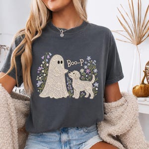 Könnte beinhalten: Ein anthrazitfarbenes T-Shirt mit Rundhalsausschnitt und einem Grafikdruck eines geisterhaften Wesens und eines Hundes in Spitzenoptik, umgeben von bunten Wildblumen. Der Text "Boo-P" steht in brauner Serifenschrift zwischen den Figuren.