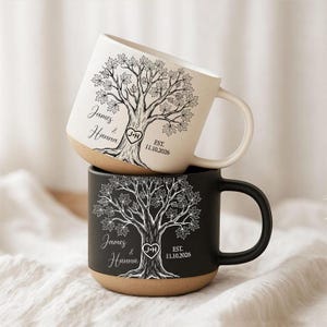 Può includere: Due tazze in ceramica, una color crema e una nera, ciascuna con un dettagliato disegno al tratto di un albero. Il design include i nomi James & Hanna, un cuore con le iniziali J+H e la scritta EST. 11.10.2026 stampata sul davanti.