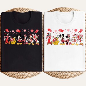 Pu&ograve; includere: T-shirt nere e bianche con un design Disney per San Valentino. La grafica presenta Topolino, Minnie, Paperino, Paperina, Pippo e Pluto, che tengono regali e palloncini a forma di cuore. Il design &egrave; su uno sfondo pieno di cuori.