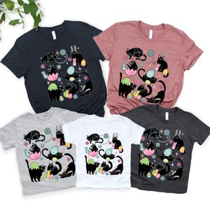 Könnte beinhalten: Verschiedene kurzärmelige T-Shirts in Marineblau, Rosa, Grau, Weiß und Anthrazit. Jedes Shirt zeigt eine Grafik mit schwarzen Katzen, Ostereiern, Hasen und Wollknäueln. Geeignet für den Alltag.