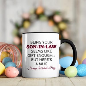 Könnte beinhalten: Eine weiße Keramik-Tasse mit schwarzem Henkel und dem Text "But First, Coffee" in schwarzer, handschriftlicher Schrift. Die Tasse steht auf einer weißen Oberfläche.