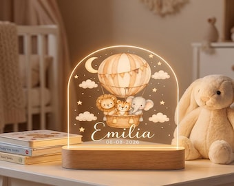 Peut inclure: Une veilleuse en acrylique en forme d'arche sur socle en bois, ornée d'une illustration de montgolfière transportant un lion, un tigre et un éléphant. Le motif inclut des étoiles, des nuages, un croissant de lune et le texte Emilia 08-08-2026.