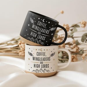Puede incluir: Dos tazas de cerámica con asa, una de color negro mate y otra de color blanco roto, cada una con el texto "COFFEE, WINGLEADERS AND HIGH LORDS" en una fuente estilizada, acompañadas de ilustraciones gráficas de alas, estrellas, una luna creciente y picos de montaña.