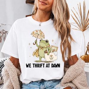 Puede incluir: Una camiseta blanca de manga corta con el gráfico de una rana verde que sostiene un paraguas floral y una bolsa de tela con un estampado de setas. La rana lleva un pañuelo estampado. Debajo de la ilustración, el texto dice WE THRIFT AT DAWN en negro.