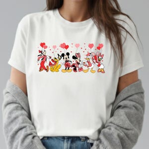 Pu&ograve; includere: T-shirt bianco con una grafica di personaggi Disney, tra cui Topolino e Minnie, che tengono palloncini a forma di cuore. I personaggi sono vestiti con abiti di San Valentino, con accenti rossi e rosa. Il disegno &egrave; centrato sulla maglietta.