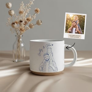 Könnte beinhalten: Weißer Keramikbecher mit braunem Boden, mit einer blauen Skizze eines Vaters, der ein Kind auf seinen Schultern trägt. Der Becher trägt die Worte "Daddy & Chloe" in blauer Schrift. Ein kleines Foto derselben Szene ist ebenfalls enthalten.