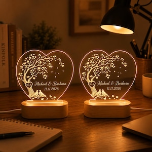 Può includere: Due luci notturne a LED in acrilico a forma di cuore con base in legno. Il design presenta un albero con foglie a forma di cuore, due uccelli e il testo inciso "Micheal & Barbara" con la data "11.11.2026" e un piccolo cuore con "M&B".