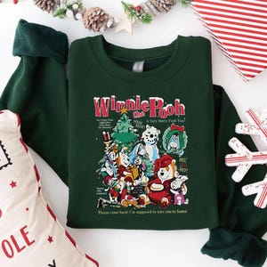 Könnte beinhalten: Dunkelgrünes Sweatshirt mit einem farbenfrohen Grafikdesign von Winnie-the-Pooh-Charakteren, die Weihnachten feiern. Die Grafik enthält den Text "Winnie the Pooh" und "A Very Merry Pooh Year". Die Ärmel sind dunkelgrün.