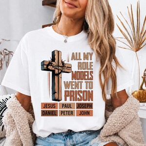 Könnte beinhalten: Ein weißes T-Shirt mit einer Grafik eines Holzkreuzes mit Dornenkrone. Der Text lautet ALL MY ROLE MODELS WENT TO PRISON, gefolgt von einem Raster mit den Namen JESUS, PAUL, JOSEPH, DANIEL, PETER und JOHN in brauner und schwarzer Schrift.
