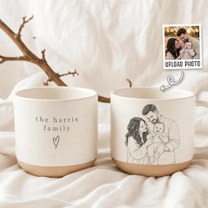 Könnte beinhalten: Zwei cremeweiße Keramiktassen mit gesprenkelter Oberfläche und beigem Boden. Eine Tasse zeigt den Text "the harris family" über einem kleinen Herzumriss. Die andere Tasse zeigt eine schwarze Strichzeichnung einer Familie mit zwei Erwachsenen und einem Baby.