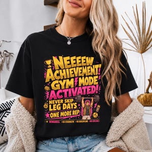 Op de afbeelding: Een zwart t-shirt met een kleurrijke afbeelding met de tekst "NEEEEW ACHIEVEMENT GYM MODE ACTIVATED! NEVER SKIP LEG DAYS ONE MORE REP" en "REWARDS: +10 STRENGTH +10 DISCIPLINE -100 EXCUSES" naast illustraties van een trofee en een gewichtheffer.