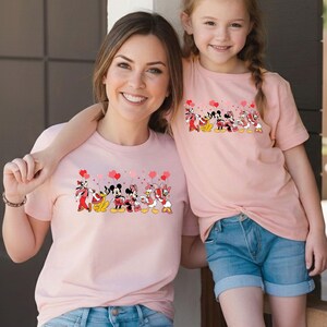 Pu&ograve; includere: Due magliette rosa abbinate con un design di personaggi Disney. Il design include Topolino, Minnie, Paperino e Paperina che tengono palloncini a forma di cuore. Le magliette sono a maniche corte e indossate da una donna e una bambina.