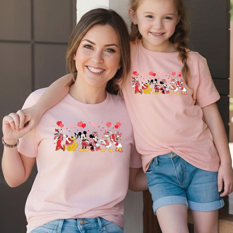Pu&ograve; includere: Due magliette rosa abbinate con un design di personaggi Disney. Il design include Topolino, Minnie, Paperino e Paperina che tengono palloncini a forma di cuore. Le magliette sono a maniche corte e indossate da una donna e una bambina.