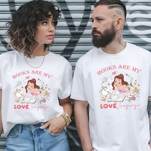 Könnte beinhalten: Weiße T-Shirts mit dem Text "BOOKS ARE MY LOVE LANGUAGE" in rosa Schrift. Die Shirts zeigen eine Cartoon-Illustration einer Frau, die ein Buch liest, umgeben von anderen Objekten. Die Shirts werden von zwei Personen getragen.