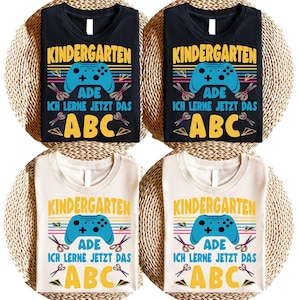 Könnte beinhalten: Vier T-Shirts, zwei schwarze und zwei cremefarbene, mit einem farbenfrohen Grafikdesign. Das Design enthält das Wort "KINDERGARTEN", einen blauen Gamecontroller, Scheren und die Buchstaben "ABC". Der Text "ADE ICH LERNE JETZT DAS" ist ebenfalls vorhanden.
