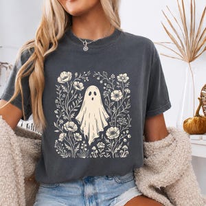 Op de afbeelding: Een antracietgrijs t-shirt met korte mouwen en een crèmekleurige grafische print van een geest in het midden, omringd door diverse botanische illustraties, waaronder bloemen, stengels en kleine paddenstoelen in een rechthoekig ontwerp.