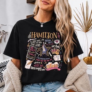 Könnte beinhalten: Ein schwarzes Kurzarm-T-Shirt mit einem Grafikdruck, der den Text "HAMILTON" und verschiedene Illustrationen wie einen Stern, eine Feder, eine Krone, eine Gitarre und Bierkrüge sowie Zitate wie "Who lives, who dies, who tells your story." zeigt.