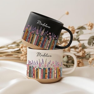 Könnte beinhalten: Zwei Keramiktassen, eine schwarz und eine cremefarben, jeweils mit einer Reihe bunter Buchrücken, die mit Lavendel und rosa Blumen verziert sind. Der Name Madeline ist in Schreibschrift auf beiden Tassen über dem Buchmotiv aufgedruckt.