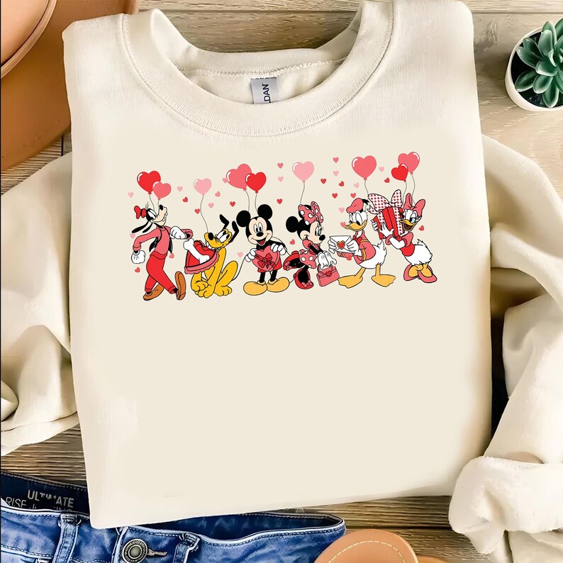 Pu&ograve; includere: Felpa color crema con una grafica dei personaggi Disney, tra cui Topolino e Minnie, Pippo, Pluto, Paperino e Paperina, che tengono palloncini a forma di cuore. I personaggi sono vestiti con abiti di San Valentino.