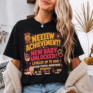Könnte beinhalten: Ein schwarzes Kurzarm-T-Shirt mit einer bunten Grafik und dem Text "NEEEEW ACHIEVEMENT! NEW BABY UNLOCKED! LEVELED UP TO DAD MISSION STARTED: PARENTHOOD REWARDS: +10 PATIENCE +1 WISDOM -100 SLEEP" sowie einer Baby-Illustration.