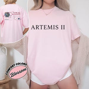 Op de afbeelding: Een lichtroze t-shirt met de tekst "ARTEMIS II" in het zwart. De achterkant van de shirt heeft een afbeelding met de tekst "FOR ALL HUMANITY" en een diagram van het zonnestelsel. Het shirt is gelabeld "COMFORT COLORS Blossom".