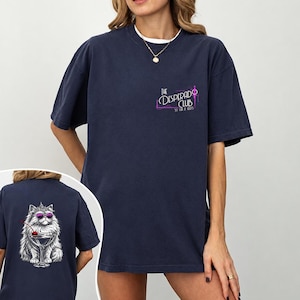 Könnte beinhalten: Ein marineblaues T-Shirt mit einer kleinen Grafik auf der linken Brust, auf der The Desperado Club steht. Die Rückseite des T-Shirts zeigt eine große Illustration einer flauschigen weißen Katze mit violetter Sonnenbrille in einem Martiniglas.
